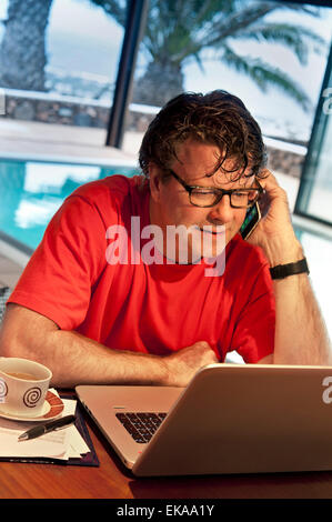 Mature man using his iPhone 6 smartphone and laptop computer in luxury vacation villa hotel interior pool room at dusk Stock Photo
