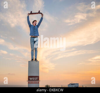 Portrait of young man standing on pillar holding skateboard above head Stock Photo