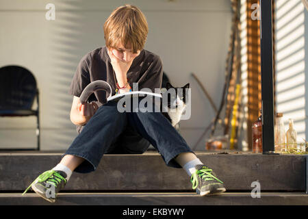 Boy sitting on step reading book, cat peering from behind Stock Photo