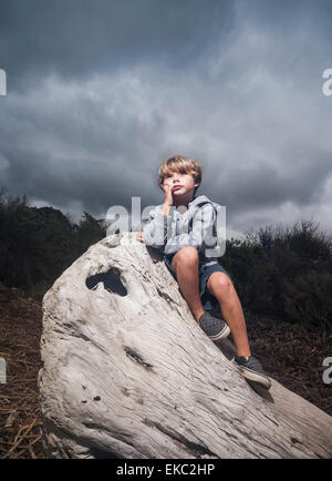 Young boy sitting on tree stump against stormy sky Stock Photo