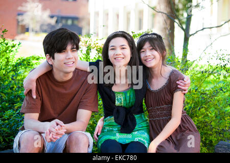 Three brother and sisters sitting outdoors on log, smiling Stock Photo