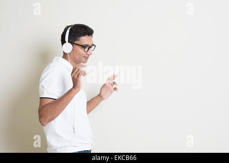 Portrait of handsome Indian guy listening to music with headphones, standing on plain background with shadow, copy space at side Stock Photo