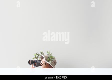 Boy looking through binoculars with some leaves and tree branches stuck on his safari hat Stock Photo