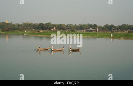 Three fishing boats glide on the placid waters of a lake in Myanmar on the shoreline people are working in the fields Stock Photo