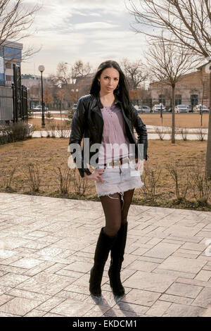 Full figure portrait of beautiful young woman in the park at fall standing alone in short skirt and black jacket Stock Photo