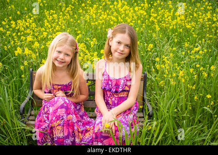 Two sisters sit together on a bench in a field of yellow flowers. Stock Photo