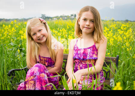 Two sisters sit together on a bench in a field of yellow flowers. Stock Photo