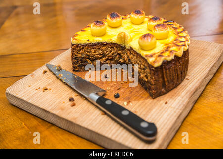 Traditional Easter simnel cake with marzipan topping and balls cut with portion removed on a wooden chopping board with a large, sharp kitchen knife Stock Photo