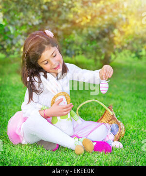 Cute little girl playing traditional Easter game, hunt for colored eggs, having fun in fresh green garden with cute little bunny Stock Photo