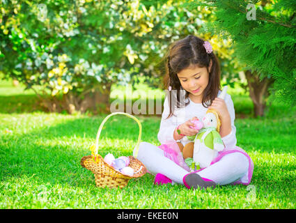 Cute little girl playing Easter game, find colorful eggs and fed them a bunny toy, having fun in fresh green garden Stock Photo
