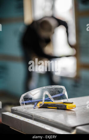 Protective Glasses and Utility Knife on a Table Saw with a worker in background Stock Photo