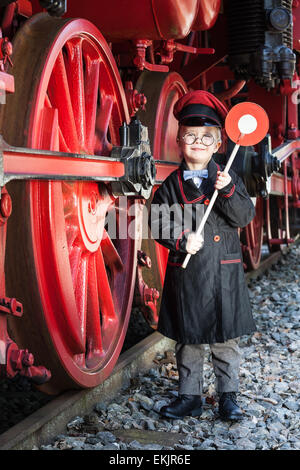 Little child boy as nostalgic railroad conductor with cap and signaling disk beside large wheels of a steam locomotive Stock Photo