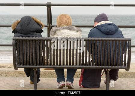 Three tourists enjoying their holiday sat on bench looking out to sea on cold overcast spring day in Looe, Cornwall, England Stock Photo