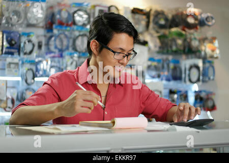 Young asian entrepreneur owning a computer shop and checking bills and invoice with calculator, doing budget and reviewing expen Stock Photo
