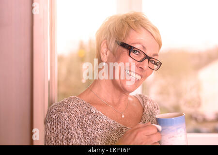 Vivacious attractive blond middle-aged woman wearing glasses standing drinking coffee and glancing sideways at the camera Stock Photo