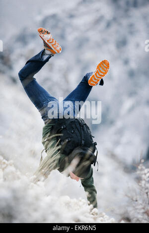 A hiker in the mountains with a rucksack, doing a handstand on a snowy slope. Stock Photo