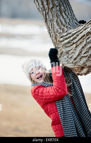 A young girl in a red jacket, and long scarf, gripping a tree trunk. Stock Photo