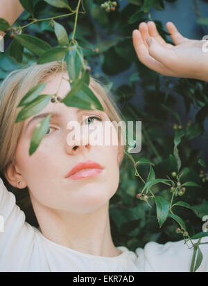 A blond haired woman looking wistfully through a vine with green leaves. Stock Photo
