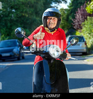 Smiling old lady on a scooter stopped in the street facing the camera giving a thumbs up of approval to show her enjoyment of th Stock Photo