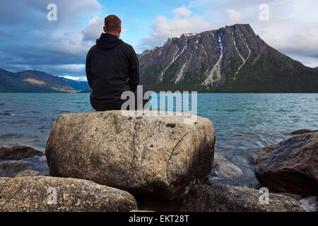 Man sitting on a rock by Kusawa Lake with Ark Mountain in the background at dusk; Yukon, Canada Stock Photo