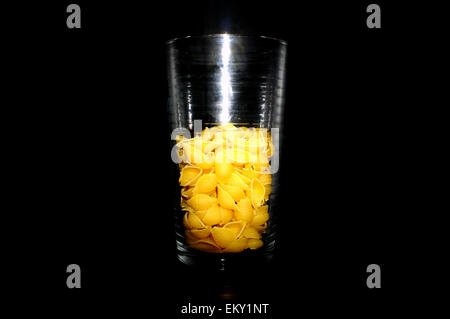 A glass half full/half empty of pasta shells against a black background. Stock Photo