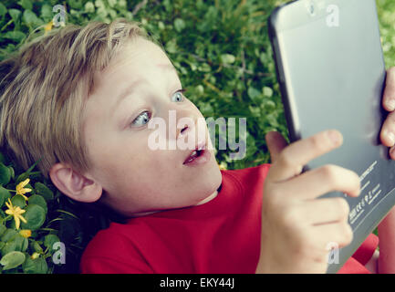 Portrait of Child blond young boy playing with a digital tablet pc computer outdoors  lying on grass lawn, facial expression Stock Photo