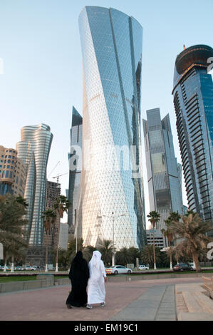 A man and woman in traditional clothes walk below skyscrapers in the city of Doha, Qatar. Stock Photo