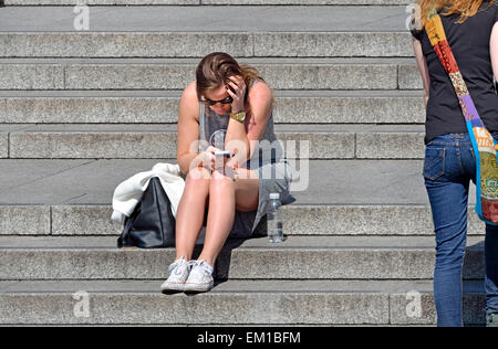 London, England, UK. Young woman sitting on the steps in Trafalgar Square, texting on her mobile phone Stock Photo