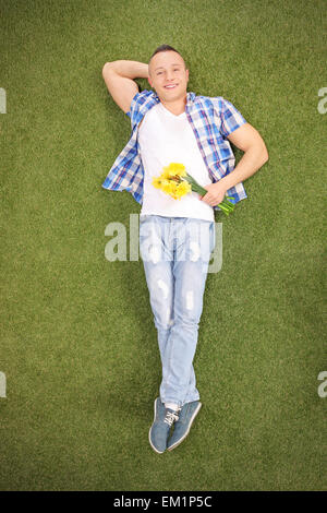 Full length vertical shot of a relaxed young guy lying on grass in a meadow, holding a bunch of yellow tulips Stock Photo