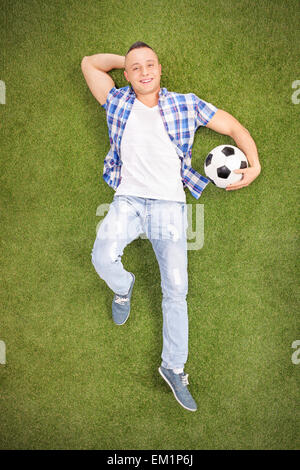 Vertical shot of a casual young man lying on a grass field, holding a football and looking at the camera Stock Photo