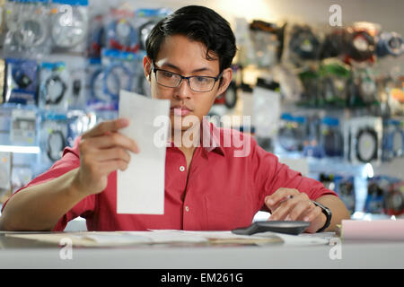 Young asian entrepreneur owning a computer shop and checking bills and invoice with calculator, doing budget and reviewing expen Stock Photo