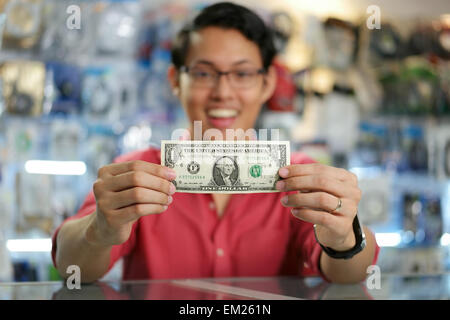 Young asian shop owner in computer store, showing his first dollar to the camera and smiling. Focus on banknote Stock Photo