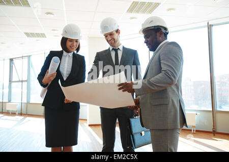 Confident architects in protective helmets discussing blueprint in office Stock Photo