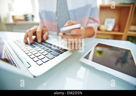 Hands of young man holding smartphone and typing on laptop Stock Photo