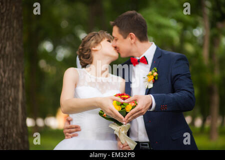 Happy bride, groom standing in green park, hands show heart - symbol of love. lovers in wedding day. happy young couple in love. Stock Photo