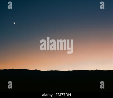 Moon rise over the silhouette of a desert landscape right after the sunset against a gradient of blue and orange sky Stock Photo