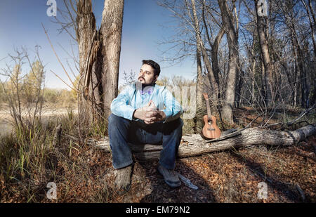 Man in blue shirt with bow tie and ukulele on the tree trunk in the forest
