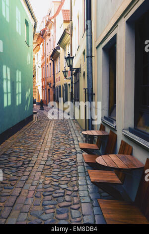 The narrow cobblestone street with reflections on the windows of colored facades and chairs outdoor cafes in Riga on a sunny day Stock Photo