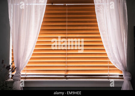 A window covered with closed wooden window blinds with white filmy curtains. Stock Photo