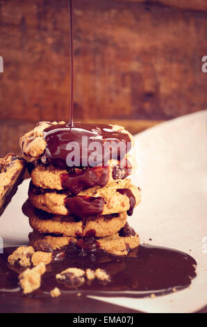 Chocolate sauce pouring over a stack of chocolate chip cookies on white curling baking paper against a dark wood background. Stock Photo