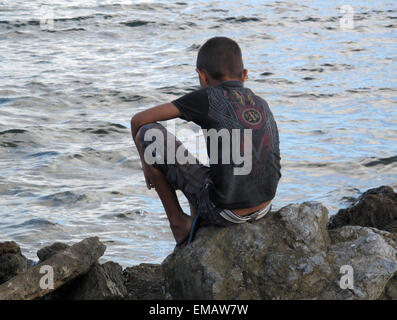 Back view of boy sitting on rock looking into water. Stock Photo