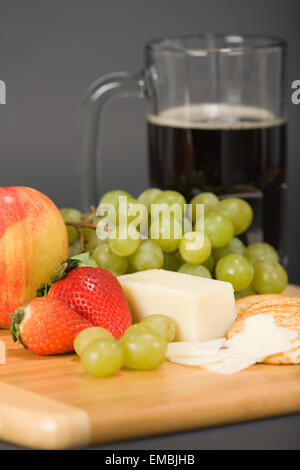 Gruyere Swiss cheese, mug of dark beer, fruit and whole wheat water crackers on a wooden cutting board Stock Photo