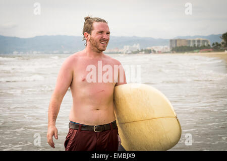 Young man carrying a surf board at the beach, looking away. Riviera Nayarit, Pacific Coast, Mexico Stock Photo