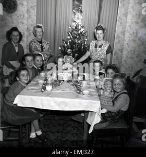 1950s, historical, A happy family xmas. Mums, grandma stand, while the exicted children, three holding dolls, sit nicely at the table prepared for their christmas meal, infront of a small cracker decorated tree. Stock Photo