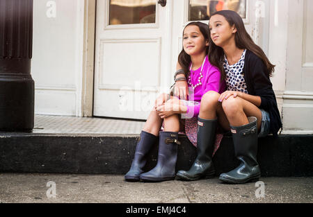 Portrait of Two Young Girls Sitting in Front of Urban Building with Older Girl Putting Arm Over Young Girl's Shoulders Stock Photo