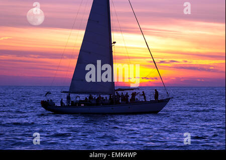 A sailboat full of people sails along the ocean at sunset as the moon rises in the background. Stock Photo