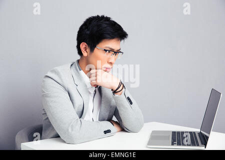 Pensive businessman sitting at the table with laptop over gray background and looking on laptop Stock Photo