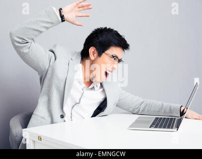 Angry young businessman in glasses sitting at the table with laptop over gray background Stock Photo