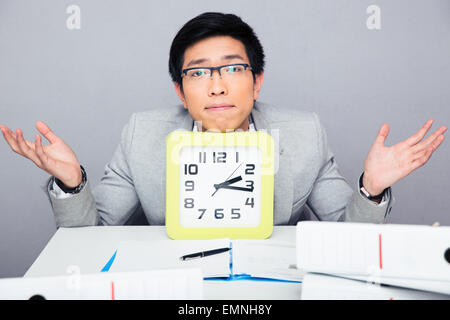 Young businessman sitting at the table with big clock and shrugging. Looking at camera Stock Photo
