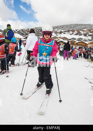 small child in skiing clothes on skis  prior to a skiing lesson with slopes  and chalets behind Stock Photo